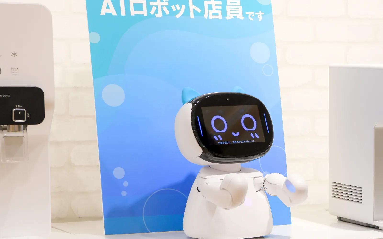 専門店の売り場で「ぼくは AI ロボット店員です」の案内板の前に立ち、画面に日本語を出して話しかける Kebbi